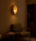 Aura Shell Wall Light glowing warmly above a cozy armchair in a minimal reading corner.