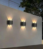 Three outdoor wall lights on a white wall with a fence and trees in the background.