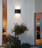 Modern outdoor wall light fixture on a building exterior with plants and glass doors.