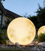 Glowing spherical outdoor light fixture on a pebble path with a house and trees in the background.