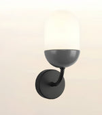 A modern wall-mounted sconce with a matte black circular backplate, curved arm, and bowl-shaped shade, paired with a frosted white dome glass shade, shown against a light beige background.