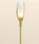 A modern tulip-shaped wall sconce with a warm glowing light bulb inside a clear glass petal-shaped shade, mounted on a brushed gold metal stem against a solid beige background.