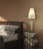 A modern wall-mounted sconce with a white tapered shade, clear glass cone, and gold-toned metal arm, lit and mounted on a warm brown bedroom wall beside a dark wood bed and patterned side table.