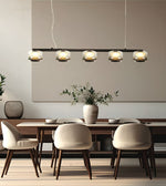 A modern 5-light linear pendant chandelier with clear glass shades and warm metallic accents, hanging over a dining table with cream upholstered chairs in a minimalist dining room.