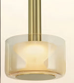 A close-up of a modern pendant light with a brushed brass cylindrical stem and a clear glass shade that glows warm yellow when illuminated, set against a plain off-white background.
