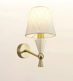 A modern wall-mounted sconce with a tapered off-white fabric shade, a white geometric cone detail, and a brushed gold metal arm and wall plate, shown against a plain light background.