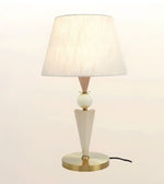 A modern table lamp with a tapered off-white fabric shade, a white spherical accent, a geometric off-white base, and a round gold metal base, shown against a plain light background.