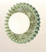 A circular decorative wall mirror with a textured, scalloped outer frame made of light green resin and embedded small pebbles, set against a plain off-white background.