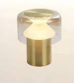 A modern table lamp with a brushed brass cylindrical base, a frosted white conical shade, and a clear glass dome top with a brushed brass lid, shown against a light beige background.