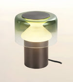A modern table lamp with a translucent smoky green glass shade, a glowing warm white frosted center, and a matte dark bronze cylindrical base on a light beige background.