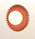 A circular wall sconce with a fluted, sunburst-like outer rim in terracotta orange-brown, featuring a large white illuminated center on a light beige background.