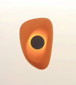 An abstract sculptural wall light or decorative object with a rounded organic shape in orange and yellow, featuring a dark circular center, shown against a plain light background.