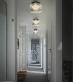 Three modern flush-mount drum ceiling lights with brushed silver metal tops and white shades, illuminated in a hallway with white walls and a hanging chair in the distance.