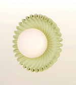 A modern wall-mounted light fixture with a textured, fluted circular shade in a pale sage green and a glowing frosted white globe bulb, shown against a soft off-white background.