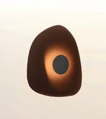 An abstract organic-shaped wall sconce or decorative object with a dark brown outer surface, a warm copper-gold illuminated center, and a dark circular opening, shown against a soft off-white background.