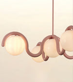 A modern pendant light fixture with a wavy, flowing frame in a muted dusty rose color and three frosted glass globes, shown against a plain off-white background.