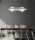 A modern pendant light with a white curved shade and black frame hangs above a wooden dining table with two chairs in a minimalist room with a textured gray wall.