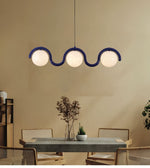 A modern three-light wavy pendant chandelier with frosted white globe shades and a dark blue frame, hanging above a dining table in a warm beige room.