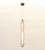A modern vertical pendant light with a brushed brass canopy and cord, featuring two stacked cylindrical glass shades with a warm glowing light inside, set against a plain off-white background.