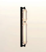 A modern vertical wall sconce light fixture with two illuminated cylindrical glass shades and a matte black metal frame, shown against a plain off-white background.