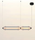 A modern two-light linear pendant lamp with clear cylindrical glass shades and warm white LED tubes, suspended by thin black cords from a black ceiling canopy against a plain off-white background.