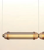 A modern cylindrical pendant light fixture with a brushed gold metal frame, textured end caps, and a ribbed clear glass shade, suspended against a plain off-white background.