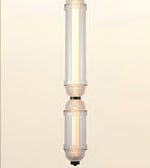 A modern vertical pendant light fixture with two stacked cylindrical glass shades, featuring a warm glowing light inside and textured metallic accents near the middle, set against a solid cream background.