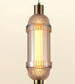 A hanging pendant light with a warm glowing cylindrical glass shade, featuring textured cut-glass details at the top and bottom and brushed brass metal accents.