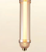 A modern cylindrical pendant light fixture with a warm glowing light inside a clear glass shade, accented by polished gold-toned metal rings and a textured glass finial at the bottom.