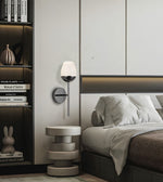 A modern wall-mounted sconce with a white frosted glass shade and a dark metal base, mounted on a bedroom wall next to a bed and a small round side table.