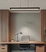 A modern black linear LED pendant light hanging above a minimalist home office desk setup with a wooden slat headboard, desk, chair, and lamp.
