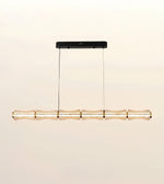 A modern linear pendant light with a textured, translucent bamboo-inspired shade and gold-toned accents, suspended from a slim black ceiling canopy against a warm off-white background.