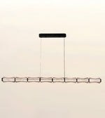 A modern linear chandelier with a textured metallic silver horizontal bar suspended by two thin cables from a black rectangular ceiling mount, shown against a plain light beige background.