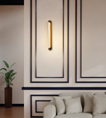 A modern vertical wall sconce with a warm glowing cylindrical light tube and brass-toned end caps, mounted on a beige wall with dark decorative panel lines above a light sofa.