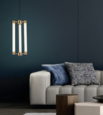 A modern three-light linear pendant chandelier with vertical frosted glass tubes and brushed brass hardware, hanging in a contemporary living room above a light gray sofa with blue and black patterned pillows against a dark navy wall.