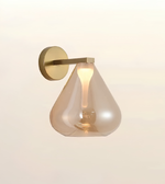 A modern wall-mounted sconce with a brushed brass metal wall plate and arm, featuring a teardrop-shaped smoked glass shade that glows warm white when lit, shown against a plain off-white background.
