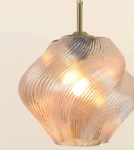 A modern pendant light with a ribbed, swirling clear glass shade that glows warm yellow when lit, suspended from a brass-colored rod against a light beige background.