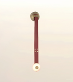 A minimalist wall-mounted pendant light with a circular gold-toned ceiling mount, a long vertical burgundy leather strap, and a glowing warm white spherical shade at the bottom, set against a soft cream background.