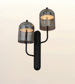 A modern double-light wall sconce with a matte black cylindrical body and two adjustable arms, each topped with a smoked translucent glass dome shade. The lights are turned on, casting a warm yellow glow, set against a plain off-white background.
