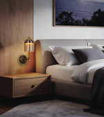 A modern bedroom scene featuring a bed with white and gray bedding, a matching wood nightstand, and a wall-mounted brass and glass bedside lamp. A wood-paneled accent wall and a framed landscape print are visible in the background.