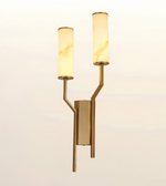 A modern double-arm wall sconce with a brushed brass/gold metal frame and two cylindrical warm-white illuminated glass shades, shown against a plain light background.