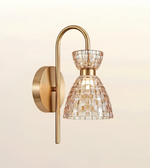 A modern wall-mounted light fixture with a curved brass arm, a round wall plate, and a textured mosaic glass shade in warm gold and clear tones, shown against a soft beige background.
