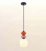 A modern pendant light fixture with a black metal rod, a round black ceiling canopy, and three stacked glass beads in orange, pink, and white, with the bottom shade glowing warm white.