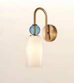 A modern wall-mounted light fixture with a curved brass arm, a round blue glass bead accent, and a frosted white glass shade, shown against a plain off-white background.