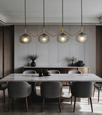 A modern dining room featuring a white marble dining table surrounded by gray upholstered chairs, under four textured glass pendant lights with gold-toned zigzag metal frames.