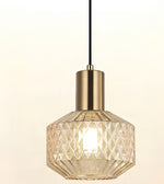 A modern pendant light with a faceted clear glass shade and a brushed brass cylindrical neck, suspended by a black cord against a warm off-white background.