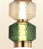 A close-up of a modern pendant light fixture featuring a faceted green glass shade below a clear textured glass shade, both illuminated with a warm yellow glow and accented by gold-toned metal rings.