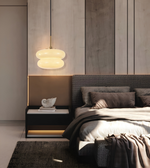 A modern bedroom interior featuring a bed with neutral-toned bedding, a dark upholstered headboard, a black bedside table with a white shell sculpture, a glowing layered pendant lamp, and a wood-paneled wall background.