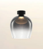 A modern pendant light fixture with a rounded, bulb-shaped transparent glass shade that fades from smoky gray at the top to clear at the bottom, topped with a matte black cap, shown against a light beige gradient background.