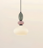 A modern pendant light with a ribbed, bell-shaped frosted white glass shade, accented by a dusty pink glass ring and a muted sage green glass bead, suspended by a thin black cord against a warm off-white background.